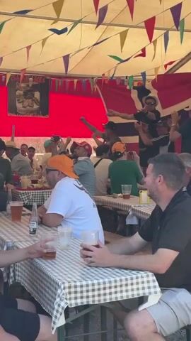 The video shows a crowded outdoor event, likely a festival or party, with a tent providing shade. Colorful pennants are strung across the tent's ceiling. A large banner with the word "Oktoberfest" is prominently displayed, featuring images of beer bottles. People are seated at long tables covered with checkered tablecloths, with drinks, including beer, on the tables. Some individuals are standing and appear to be dancing or interacting enthusiastically. The atmosphere seems lively and social. The camera pans across the scene, capturing various groups of people and the festive decorations.