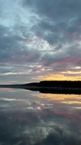 The video shows a serene lake at dawn or dusk. The sky is filled with dramatic clouds painted in hues of gray, pink, and yellow, reflecting beautifully on the still surface of the water. A dark, treeline forms the distant horizon, with a faint suggestion of buildings or structures along its edge. The overall atmosphere is peaceful and tranquil, with the camera slowly panning across the scene, capturing the gentle play of light and color on the water and in the sky. There are no people or discernible objects in motion, creating a sense of stillness and natural beauty.