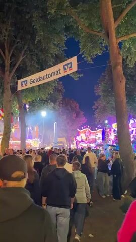 The video shows a crowded outdoor event at night, likely a fair or festival. People are walking around, some are standing in groups, and the atmosphere is lively. In the background, there are illuminated amusement park rides and tents, with colorful lights and some smoke or fog. A banner reading "Geldautomat" (ATM) with a bank logo is visible hanging between trees, indicating the presence of a banking service. The sky is dark blue, suggesting it's late evening or night. The trees overhead are lush and green, providing a natural canopy. The camera is moving, capturing different sections of the event as it pans. The overall impression is a bustling public gathering with entertainment and amenities.