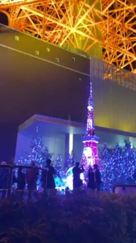 The video shows the Tokyo Tower at night, illuminated in orange. At the base of the tower, a small replica tower stands illuminated in purple, pink and blue. Several trees with blue lights are also visible, and a group of people stand behind a railing looking at the displays. The camera pans upwards, showing the full height of the tower.