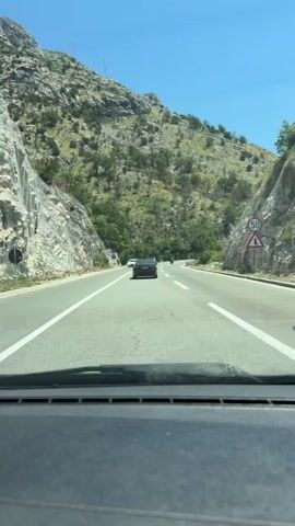 The video shows a view from inside a car driving on a paved road. The road is surrounded by rocky, mountainous terrain with sparse vegetation. In front of the car, there are two other vehicles: a white car in the left lane and a dark-colored car in the right lane. Further ahead, there appears to be a motorcycle in the distance on the right side of the road. The sky is clear and blue. The perspective suggests the camera is positioned on the dashboard or windshield of the car.