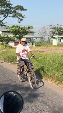 The video captures a man riding an old bicycle on a paved road. He is wearing a white t-shirt, dark shorts, and a light-colored baseball cap. The man appears to be of older age and is looking towards the camera with a slight smile. The bicycle has a basket attached to the handlebars and appears to be well-used. The road is flanked by tall green grass on the right side. In the background, there are buildings, trees, and a clear blue sky, suggesting a rural or suburban setting during the daytime. The footage seems to be taken from another vehicle, as a portion of a mirror is visible in the lower left corner of the frame.
