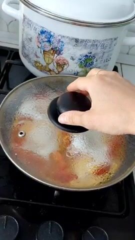 A hand with a light skin tone is shown lifting the lid of a frying pan on a stovetop. The pan contains four eggs cracked into it, mixed with chopped red tomatoes. The eggs are in the process of being cooked. A large white pot with a floral design is visible behind the frying pan. The stovetop is black. The lighting appears to be from above.