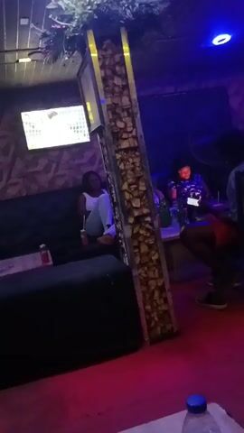 The video depicts the interior of what appears to be a lounge or bar with dim, colorful lighting. Three individuals are visible: one woman is seated on a dark-colored couch, a second woman is seated at a table with beverages, and a man is partially visible, also near the table. A large, decorative pillar stands prominently in the center, adorned with greenery on top and what appears to be textured stones or panels along its height. A television screen is mounted on the wall displaying a video or broadcast. A man wearing shorts walks in front of the camera. The floor is covered with a red carpet.
