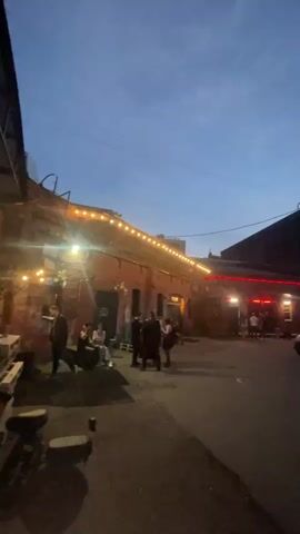 The video shows a dimly lit outdoor scene, possibly an alleyway or courtyard, at dusk. The sky is a deep blue, transitioning into night. Brick buildings line the sides of the scene, illuminated by strings of warm-toned lights draped along one building and red neon lights above an entrance on another. Several people are visible, some standing in small groups, others walking. One group is seated on steps. Cars are parked along the right side of the scene. The overall atmosphere appears to be social and relaxed, perhaps outside a bar or venue.