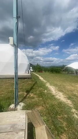 The video opens with a close-up view of a wooden structure and a blue pole with wires. The camera then pans to the right, revealing a white geodesic dome on the left side of the frame, with a wooden deck in front of it. A dirt path leads away from the dome into a grassy field dotted with trees. In the distance, a white tent structure with a curved roof is visible. The sky is a mix of blue and white clouds. As the camera continues to pan, more trees and a hilly landscape come into view. A small wooden structure with a dark roof is seen on the right side of the frame. The scene appears to be an outdoor recreational area or glamping site.