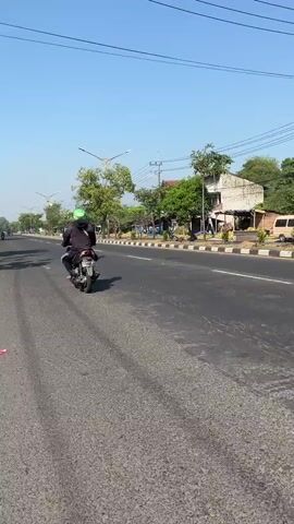 A person rides a scooter down a paved road on a clear, sunny day. The rider wears a black jacket and a bright green helmet. The scooter has a license plate that reads "AD 4836 CKB". 

The road is lined with trees and a sidewalk. Buildings are visible on the side of the road, and one appears to be a small shop or restaurant. In the background, other vehicles can be seen, including another scooter and a van. The sky is a vibrant blue with no clouds. The overall impression is a bright, warm day in a suburban or rural area. 

The scene appears to be a typical day on a road in Ketapang, Indonesia. The time of day is likely late morning or early afternoon, given the bright sunlight and clear sky. The rider is simply commuting, with no indication of any special event or activity. The general atmosphere is calm and ordinary.