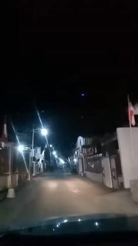The video shows a dark, empty street lined with houses. Streetlights illuminate the road and the fronts of the houses, casting long shadows. The perspective suggests the video is being recorded from a moving vehicle. The houses appear to be residential, with some having visible decorations or architectural details. The sky is black, indicating it is nighttime.