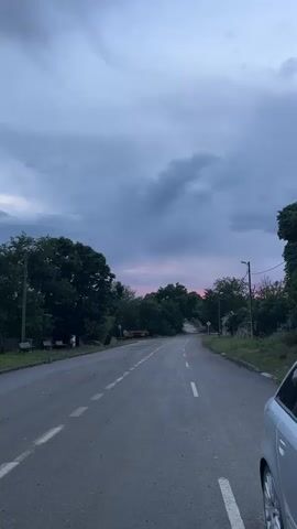 The video captures a street view from a moving perspective in Kosten, Bulgaria, under an overcast and cloudy sky. At the beginning, a two-lane asphalt road with white dashed lines is visible, curving gently to the right. The horizon in the distance shows a subtle reddish-pink glow, suggesting either dawn or dusk. Lush green trees line both sides of the road. On the left, a utility pole stands near two park benches. A silver car is partially visible on the right side of the frame. As the camera moves, it pans past a white, single-story house with a red-tiled roof and a black trash can on the left side of the road, revealing more trees and a general residential or semi-rural setting. No specific activity or visible text is present.