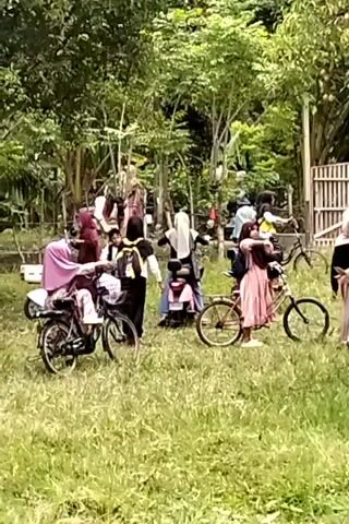 A group of people, mostly women wearing hijabs, are gathered outdoors in a grassy area with trees in the background. Several people are on or near bicycles and a scooter. The individuals appear to be interacting with each other, some are seated on the vehicles, while others are standing. The scene suggests a casual gathering or outing.