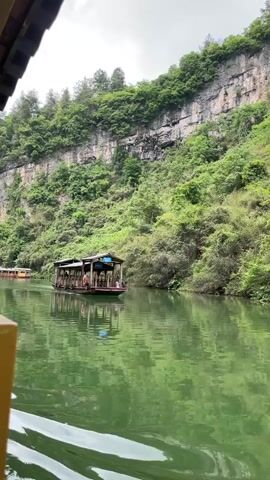 The video shows a small wooden boat on a wide, green river. The boat is covered and has railings around the edges. There are a few people on the boat, though they're not clearly visible. The river is surrounded by steep, rocky cliffs covered in lush green trees and vegetation. The sky is overcast and gray. The video is taken from another boat, with part of its roof visible in the upper left corner and part of its side visible in the lower left corner. The water is calm, and the boat is moving slowly.
