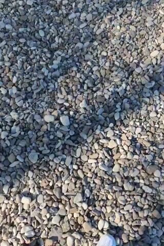The video shows a first-person perspective of someone walking on a gravel path. The gravel is composed of small, rounded stones in various shades of gray, white, and brown. The person is wearing white slip-on shoes and dark shorts. The sun is casting shadows on the gravel, indicating it is daytime. The camera is pointed downwards, focusing on the ground and the person's feet as they walk.