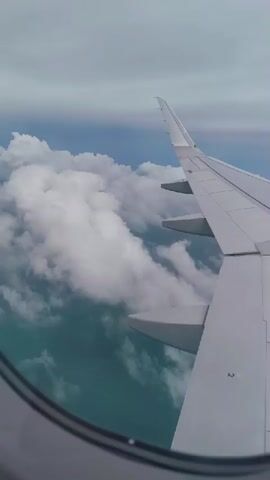 The video shows a view from an airplane window. The airplane's wing is visible on the right side of the frame. Below the wing, clouds and a turquoise body of water can be seen. The sky is overcast.