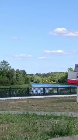 A wide shot shows a monument in the shape of a grey and red ship, positioned on a white pedestal. The monument is situated outdoors with a body of water and lush green trees in the background. A black metal fence runs along the edge of the water. The sky is blue with scattered white clouds. In the foreground, there is green grass and a paved path. Two bicycles are parked near the fence.