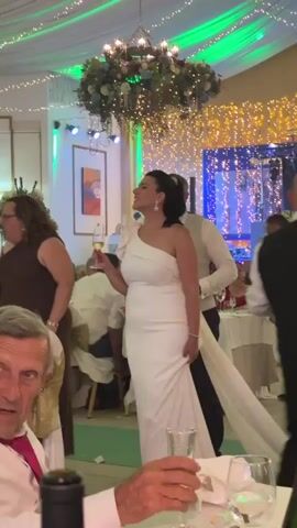 A wedding reception is in progress. A woman in a white, one-shoulder wedding dress holding a champagne flute raises it and smiles. A man in a light-colored vest and shirt stands behind her. A woman in a brown dress stands to the left. Other guests are seated at tables in the background. The room is decorated with white draping, string lights, and a floral arrangement hanging from the ceiling.