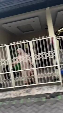 The media file shows a person wearing a hijab and a light-colored garment, walking behind a white metal gate. The gate has vertical bars and a decorative pattern of circles. The person is blurred, suggesting movement or a low-quality recording. Behind the gate, a house is visible with a covered porch and a ceiling. The ground in front of the gate appears to be paved, with a darker strip running along the bottom edge, possibly a road or sidewalk. The scene seems to be outdoors, during the daytime, with neutral weather conditions. The location is identified as Ketegan, Indonesia. No specific emotions or activities are clearly discernible due to the blurriness and lack of detail. There is no visible text in the media file.
