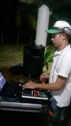 A man in a white shirt and green-brimmed baseball cap is DJing on a patio at night. He’s using a laptop, a mixing board, and a speaker. The camera pans out to show a tiled patio area with wooden chairs and tables. Several people are seated, standing, or looking at their phones. The area is illuminated by hanging lights and surrounded by greenery.