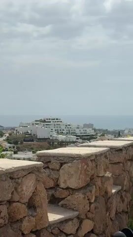The video shows a landscape scene with white buildings in the background, possibly hotels or apartments, overlooking the ocean. The sky is cloudy. In the foreground, there is a stone wall with plants growing on it. The camera pans to the right, revealing a neatly trimmed, spherical bush. Further to the right, the scene shifts to show two people walking away from the camera, with the ocean and sky in the background. Another person is visible to the left, standing and facing away. The video ends with a view of the ocean under a cloudy sky, with more greenery and stone structures in the foreground.
