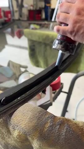 A close-up shot shows a person using a caulking gun to apply sealant around the edge of a piece of glass, presumably a window. The glass is resting on a stand covered with towels. The person's hands are visible as they operate the caulking gun, applying a bead of black sealant into the crevice between the glass and the black rubber frame. The caulking gun is red with a silver nozzle, and appears well-used. In the background, the interior of a workshop is visible, with red cabinets, tools, and a ladder. The lighting is bright. An air compressor with a hose is visible in the foreground.