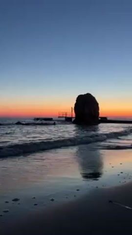 The video shows a beach at sunrise or sunset. The sky transitions from a deep blue at the top to orange and yellow near the horizon. A large rock formation stands in the sea, silhouetted against the colorful sky. A small pier or structure is visible next to the rock. Gentle waves are washing onto the sandy beach, reflecting the colors of the sky. The scene appears peaceful and serene.