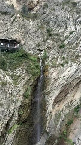 The video shows a long shot of a narrow waterfall cascading down a sheer, rocky cliff face. The cliff is composed of layered rock, with some sparse greenery growing in the crevices. On the left side of the cliff, near the top, a small wooden structure with a balcony is visible. Several people can be seen standing on the balcony. A thin wire or cable stretches across the frame from the structure towards the right side of the cliff, and another similar cable runs from the structure downwards. The water from the waterfall flows down the rocks and disappears into the lower part of the frame.