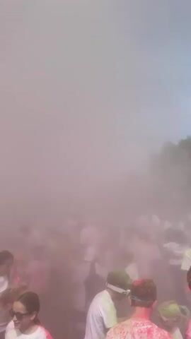 The video shows a large crowd of people participating in a color run or similar event outdoors. Most people are wearing white shirts and are covered in colorful powder, predominantly pink. There's a cloud of colored dust hanging in the air, partially obscuring the view. People are walking around, some smiling and interacting. In the background, trees and buildings are visible, along with an inflatable structure.
