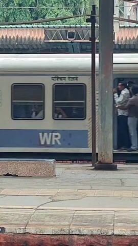 A train is stopped at a station. The doors of the train are open and several people are standing in the doorway. Some people are looking out of the train and others are talking to each other. The train is white and blue and has windows. The station platform is made of concrete.