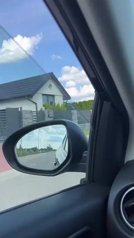 The video captures a view from inside a moving car, looking out the passenger side window and in the side mirror. The sky is blue with white clouds. In the side mirror, the road behind the car is visible, with another car in the distance. Outside the window, there is a roadside with green grass, some trees, and a tall metal electrical tower. The interior of the car, including a black door panel and an air vent, is also visible. The car appears to be driving on a paved road.