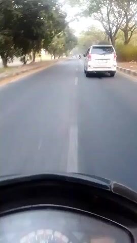The video shows a first-person perspective of someone riding a scooter on a road. The scooter's dashboard is visible in the foreground. In the middle ground, a white car is driving ahead on the same road, and there are trees lining the side of the road. The road appears to be paved and is in daylight.
