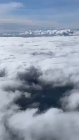The video shows a view of clouds from above, likely taken from an airplane. The clouds are dense and white, with some darker areas below. The sky is blue in the upper part of the frame. The image is static throughout the video.