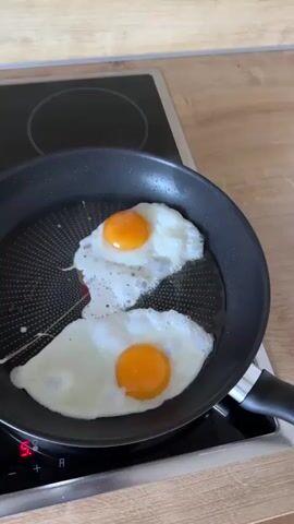 The video shows two fried eggs sizzling in a black non-stick frying pan on a black induction stovetop. The eggs are sunny-side up, with bright orange yolks and white egg whites that are beginning to brown slightly around the edges. The pan is placed on the front left burner of the stovetop, which is displaying a red indicator light, suggesting it is in use. The stovetop is set against a light-colored wooden countertop. The camera is positioned above and slightly to the side of the pan, capturing a close-up view of the cooking eggs. The overall scene depicts a common kitchen activity: preparing breakfast.