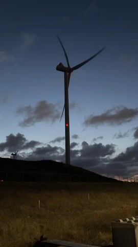 The video shows a wind turbine in the evening or at night. The sky is dark blue with some clouds. The wind turbine has three blades, and they are spinning. There are red lights on the turbine, one near the top of the tower and one higher up near the hub. There is another, smaller wind turbine in the background on the left, also with red lights. The foreground is dark and shows dry grass and a fence.