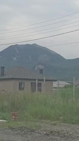 The video shows a rural scene with houses and mountains in the background. The sky is overcast. There are power lines in the foreground and a fence running across the middle of the frame. The houses have various colored roofs and are surrounded by tall grass. One house in the foreground appears to be under construction or unfinished. The overall impression is one of a quiet, undeveloped area.