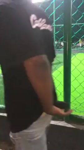The video shows a group of men playing soccer on a green indoor field. The players are wearing different colored jerseys, including light blue and green. The field is enclosed by a green net. There are a few people watching the game from outside the net. The camera is held by someone standing near the net.