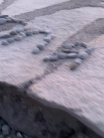 The video opens with a slightly blurry, close-up shot of small, dark grey, rounded stones arranged in a serpentine, winding pattern on a light-colored, possibly concrete or stone paved surface. Another, straighter path of similar stones is visible in the background on the same surface.

The camera then slowly pans upwards and to the right, revealing the edge of the paved surface where it meets a body of water. The water is dark blue-grey with gentle ripples. In the distance, across the water, bright lights from buildings or a settlement are visible along the shoreline.

As the camera continues to pan, it captures a wide, panoramic view of the tranquil water body. The sky above displays a beautiful gradient of colors, starting with warm orange and yellow near the horizon, transitioning through soft pinks, and fading into a light purple-blue higher up. This color palette strongly suggests either sunset or sunrise. The distant shoreline is a dark silhouette of hills or mountains, dotted with numerous bright, warm-toned lights, indicating a town or city along the water's edge. A single white buoy floats in the calm water. The water itself reflects the colors of the sky, showing subtle hues of pink, orange, and blue-grey. The overall atmosphere is peaceful and serene, depicting a beautiful twilight scene over a bay or large lake.