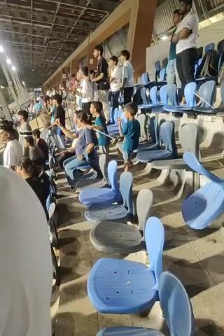 The video shows a crowd cheering from the stands at a sports stadium. Most of the crowd is facing the field and raising their hands in the air in excitement. The stadium has blue and gray plastic seating with some seats flipped up. The field is visible with athletes in blue uniforms, apparently celebrating a win. The environment appears to be outdoors under stadium lights, with urban buildings visible in the distance.