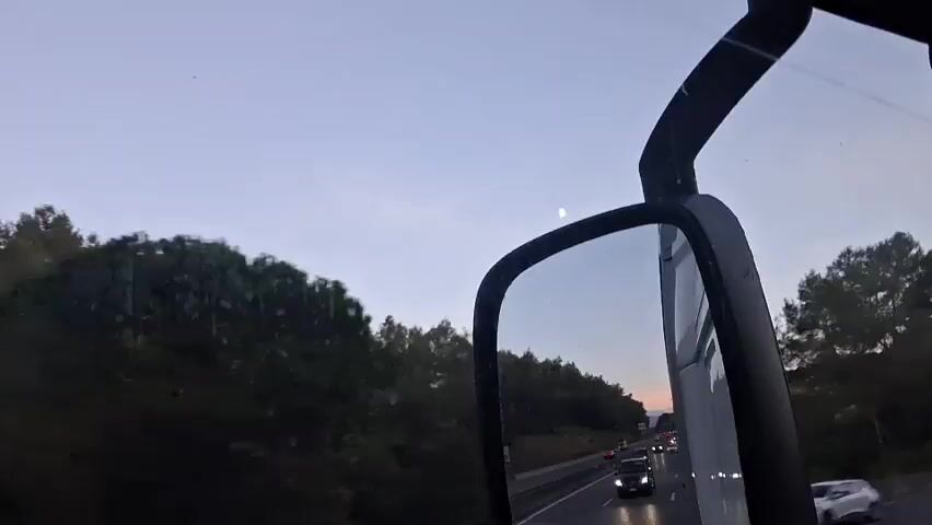 The video captures a view from the interior of a vehicle, likely a truck, looking out the passenger side window and reflecting in the side mirror. The time appears to be dusk or early dawn, with a pale blue sky and a crescent moon visible. The landscape outside is dominated by trees and foliage, suggesting a rural or semi-rural setting. The side mirror reflects a highway with several vehicles, including cars and at least one other truck, traveling in the same direction. The headlights of these vehicles are illuminated, indicating low light conditions. The overall scene conveys a sense of travel on a highway during twilight.