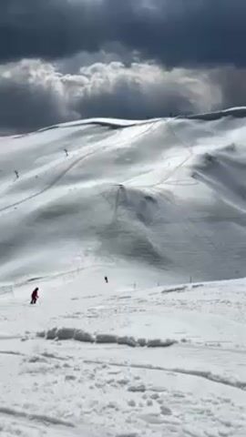 The video shows a wide shot of a snowy mountain landscape under a cloudy sky. Several skiers are visible on the slopes. The camera zooms in on one skier in the lower right of the frame, dressed in black and skiing down the hill, leaving tracks in the snow. The focus remains on this skier, showing their solitary descent.