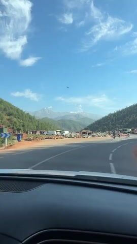 The video shows a view from inside a car as it drives on a road, presumably in Morocco, toward a mountainous area. Several motorcyclists pass by the car. The mountains are visible in the distance, covered in snow. Along the side of the road are stalls or shops selling what appears to be pottery. The sky is blue with scattered clouds. A bus is parked along the side of the road.