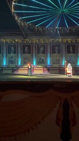 The video shows an outdoor performance staged against a backdrop resembling ancient Roman architecture with murals depicting mythological scenes. Two figures are on a dirt stage. The figure on the left is dressed in a long, dark, sequined robe and a red horned mask, holding a lit torch that emits flames. The figure on the right is dressed in a white, hooded robe and holds a staff with a glowing orb at the top. The stage is lit with spotlights, and decorative string lights hang overhead, creating a festive atmosphere. The backdrop features a frieze of sculptures and painted panels. A large, illuminated structure with radiating lights is visible in the upper portion of the frame. The overall impression is that of a theatrical or ceremonial event with a mystical or historical theme. The audio includes spoken Russian.