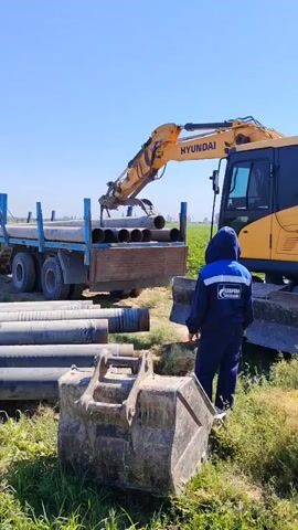 The video shows an excavator, a truck, and a worker on a bright, sunny day. The excavator is yellow and has the brand name HYUNDAI visible on its arm. It is actively lifting a long, cylindrical pipe from the truck's flatbed. The truck is blue and appears to be a flatbed trailer with several pipes already loaded on it. The worker is wearing a dark blue uniform with a hood, and a logo with the words "GAZPROM" and a blue and white emblem is visible on the back of the jacket. The worker is standing on the ground, looking towards the excavator and the truck. In the foreground, there is a large excavator bucket resting on the ground, surrounded by grass and some additional pipes of varying sizes. The environment appears to be a rural or undeveloped area with green fields and a clear blue sky.