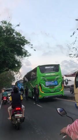 A street scene captured from behind a person on a motorcycle, with a bright pink helmet and pink headscarf visible in the foreground. Several other motorcycles are on the road, with riders. In the middle ground, a large green bus with "RODEVA" and "PUTRA TRANS" written on it is stopped in traffic. A person stands next to the bus, holding up a white flag. Other buses and vehicles are also in the traffic jam. Trees line the left side of the road, and the sky is overcast. The overall atmosphere suggests a busy, congested road during daylight hours.