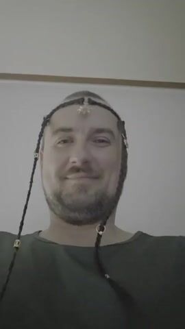 A man with a headset and braids is smiling directly at the camera. The video is a close-up shot of his face with a plain wall in the background. The man appears to be wearing a dark green shirt and has some decorations in his braided hair.
