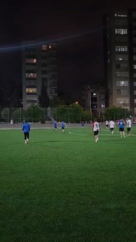 The video shows an outdoor soccer field at night. Artificial lights illuminate the green turf. Several people are playing soccer. Two tall apartment buildings are visible in the background. The sky is dark.