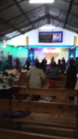 Inside a church, a group of people is seen from behind, facing the altar. The church has rows of wooden pews, and people are standing and moving around. The altar area is decorated with neon lights, crosses, and a menorah-like object. A screen hangs above the altar. People are wearing various clothes, including a yellow shirt, a red coat, and a black and white jacket. The room is lit with fluorescent lighting.