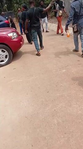 The video shows several people gathered on a dirt road next to a red car. A woman wearing a black outfit walks past the car and smiles briefly towards the camera. Other individuals are walking around, some dressed casually in jeans and shirts. One person is carrying a small bottle. The environment appears to be outdoors, possibly in a residential area or gathering space near trees and buildings.
