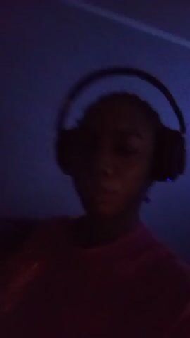The video shows a person with dark skin and hair wearing headphones and a red shirt. The person is vaping, and smoke is visible exhaling from their mouth. The background is dark blue.