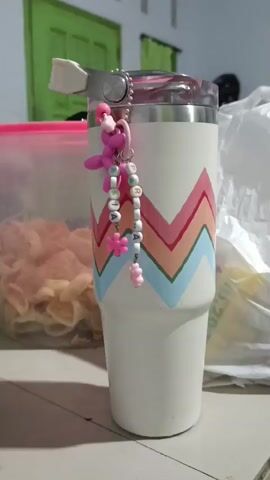 The video shows a close-up of a white tumbler with a zig-zag pattern in pink, orange, and blue. A pink balloon dog charm hangs from the top of the tumbler, along with a beaded keychain spelling out "ALIA". To the left of the tumbler is a clear plastic container filled with light-colored snack-like items. In the background, a green door is visible, and part of a white wall with light fixtures. A white plastic bag is partially visible on the right. The tumbler is placed on a surface that appears to be a table or countertop. The lighting suggests an indoor environment, possibly a home.