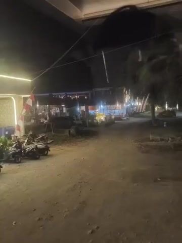 A nighttime outdoor shot shows a row of motorcycles parked on a dirt or gravel lot in front of a building. The building has a modern design with arched window openings illuminated by white string lights. A sign with vertical text is also illuminated. Plants are placed in front of the building, and some industrial barrels are visible to the right of the entrance. A red and white flag is mounted on the side of the building. The overall impression is of a business or establishment that is open late.