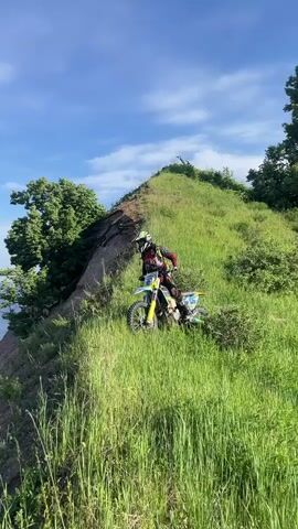 A person on a dirt bike wearing a helmet and protective gear is on a grassy hill overlooking a large body of water. The person briefly moves the dirt bike forward, then turns it and rides down the grassy slope towards the camera. The camera then pans down to show the steep, eroded hillside leading to the water, with a distant shoreline visible. The sky is blue with some clouds.